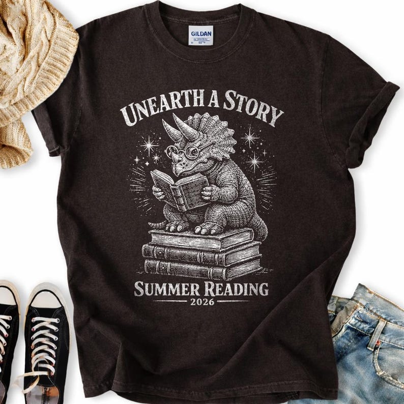 May include: A black short-sleeve t-shirt featuring a white graphic of a triceratops wearing glasses and reading a book while sitting on a stack of books. The text reads "UNEARTH A STORY" above and "SUMMER READING 2026" below the illustration.