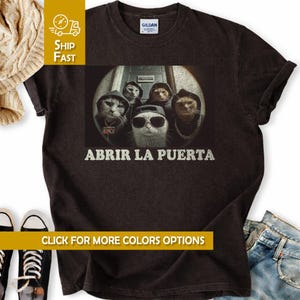 May include: Dark brown t-shirt featuring a graphic of five cats dressed in hip-hop attire, with the text "ABRIR LA PUERTA" in white. The cats are wearing hoodies, sunglasses, and chains. The shirt has a crew neck and short sleeves.