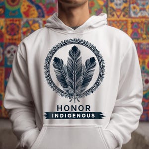 May include: White hooded sweatshirt featuring a circular design with three dark blue feathers. The design is surrounded by an ornate border. Below the design is the text "HONOR INDIGENOUS" in white letters on a dark blue banner.