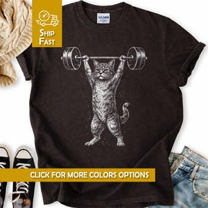 Könnte beinhalten: Dunkelgraues T-Shirt mit einer detaillierten Illustration einer Katze, die eine Langhantel über dem Kopf hebt. Die Katze ist im Vintage-Stil dargestellt, mit dem Text "CLICK FOR MORE COLORS OPTIONS" unten. Das Shirt hat auch den Text "SHIP FAST" oben links.