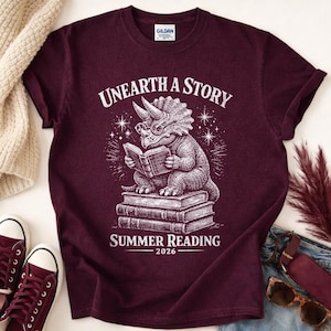 May include: A maroon short-sleeved t-shirt featuring a white graphic of a triceratops wearing glasses and reading a book while sitting on a stack of books. The text reads "UNEARTH A STORY" above and "SUMMER READING 2026" below the dinosaur.