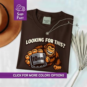 May include: A brown t-shirt with the text "LOOKING FOR THIS?" and a cartoon ape holding a 10mm socket. The shirt has a crew neck and a silver necklace. The image also includes a "Ship Fast" graphic and a "Click for more colors options" graphic.