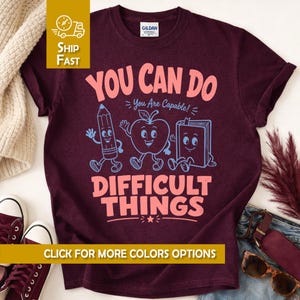 May include: A dark maroon short-sleeve t-shirt featuring a graphic print with the text "YOU CAN DO DIFFICULT THINGS" in light pink and "You Are Capable!" in light blue, accompanied by cartoon illustrations of a pencil, an apple, and a book.