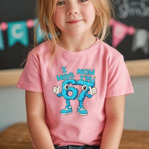 Pu&ograve; includere: Una maglietta rosa con la scritta "I WAS 6, NOW I'M 7" in carattere pixelato blu. I numeri 6 e 7 sono personificati con braccia, gambe e scarpe. La maglietta &egrave; indossata da un bambino.