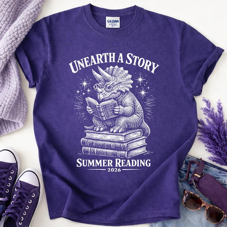 May include: A purple short-sleeve t-shirt featuring a white graphic of a triceratops wearing glasses and reading a book while sitting on a stack of books. The text reads UNEARTH A STORY and SUMMER READING 2026 in a white serif font.