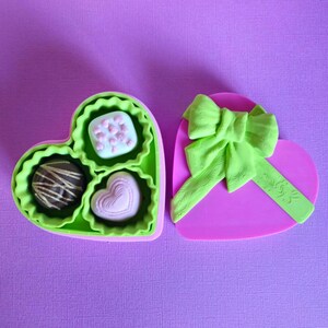 Puede incluir: Una caja de bombones en forma de corazón, rosa con lazo verde. El interior verde contiene tres bombones. El fondo es morado liso.