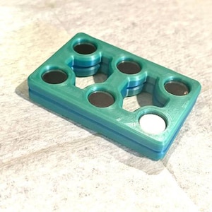 Puede incluir: Un objeto rectangular de color verde azulado y azul con seis aberturas circulares, cada una con un círculo oscuro. Una abertura tiene un círculo blanco. El objeto está hecho de material en capas.