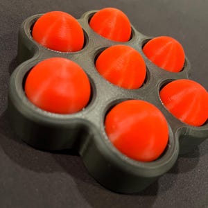May include: A black, flower-shaped holder containing seven bright orange spheres. The spheres are evenly spaced within the holder, which has a matte finish. The spheres appear to be made of a solid material.