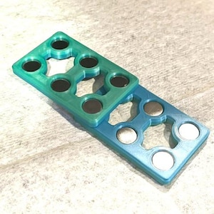 以下が含まれることがあります： ティールとブルーの色合いの2つの長方形の幾何学的なオブジェクトが積み重ねられています。それぞれに円形の切り抜きと埋め込まれた磁石があります。上のオブジェクトはティールで、下は青です。オブジェクトは明るい色の表面にあります。