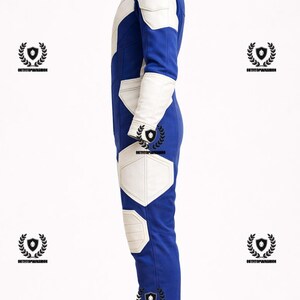 May include: Blue and white jumpsuit with a high collar. The suit features white accents on the shoulders, arms, and legs. The design includes white geometric shapes on the chest and knees. The overall style is sleek and modern.