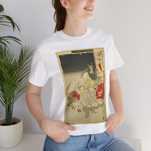 Puede incluir: Camiseta blanca con una impresión japonesa vintage en madera. La impresión representa una figura con túnicas fluidas y barba larga, sosteniendo un bastón. La obra está enmarcada por un borde rojo y dorado.