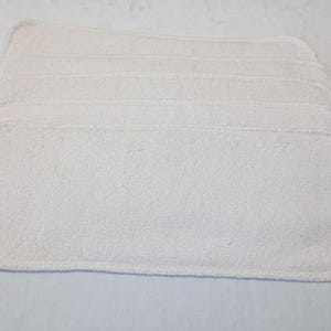 May include: Six white rectangular cloth diaper inserts are stacked. The inserts are made of a soft, absorbent material with rounded corners. They are neatly arranged, suggesting a practical, reusable baby product.