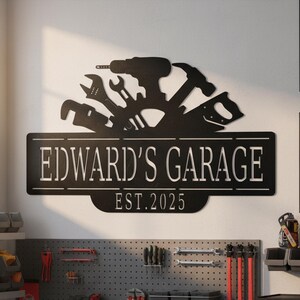 May include: Black metal garage sign with the text "EDWARD'S GARAGE" and "EST. 2025". The sign features silhouettes of tools like a drill, hammer, and wrenches. The sign is mounted on a white wall.
