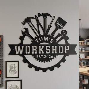 Puede incluir: Letrero de taller de metal negro con el texto "Tom's Workshop EST.2026" y siluetas de herramientas. El letrero está montado en una pared gris claro, con bocetos enmarcados y una placa perforada visible.