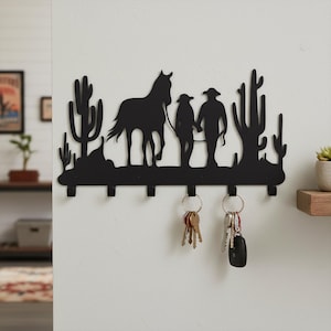 May include: Black metal key holder featuring a silhouette of a horse, two people, and cacti. The key holder has six hooks and is mounted on a white wall. Keys hang from two of the hooks.
