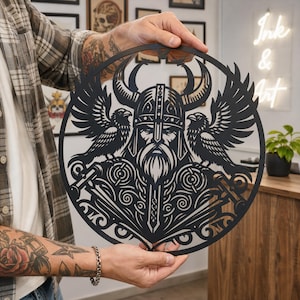 Op de afbeelding: Een cirkelvormig zwart metalen wandkunstwerk met een gedetailleerd uitgesneden ontwerp van een Vikingkrijger met een gehoornde helm, geflankeerd door twee raven met gespreide vleugels en versierd met ingewikkelde patronen.