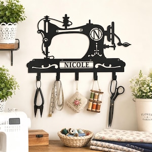 May include: A black metal wall-mounted hook rack shaped like a vintage sewing machine, featuring the name NICOLE cut out in the center. The rack has five hooks holding sewing tools including scissors, a measuring tape, and thread spools.