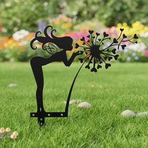 May include: Black metal garden stake featuring a silhouette of a fairy blowing on a dandelion with heart-shaped seeds. The fairy has long hair and wings. The stake is designed to be placed in a garden or lawn.