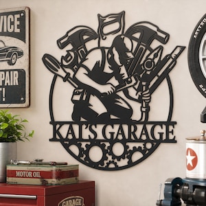 May include: A black metal wall sign featuring a silhouette of a mechanic working with tools, including a hammer, saw, and wrench. The circular design incorporates gear motifs at the bottom and the text KAI'S GARAGE in bold capital letters.
