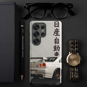 May include: A white phone case featuring a sports car design with the text "Nissan" in Japanese. The case is surrounded by a black pencil, glasses, a gold watch, and a black notebook on a dark surface.