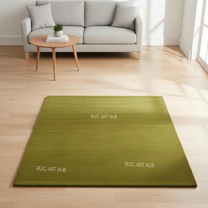 May include: A rectangular, olive green rug with a subtle gradient effect. The rug is placed on a light wood floor in a living room setting. The words "RUG ART HUB" are printed on the rug in white. A gray sofa and a wooden coffee table are in the background.