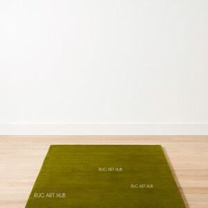 May include: A rectangular, olive green rug with a textured surface. The rug is placed on a light-colored wooden floor against a white wall. The text "RUG ART HUB" is printed on the rug.