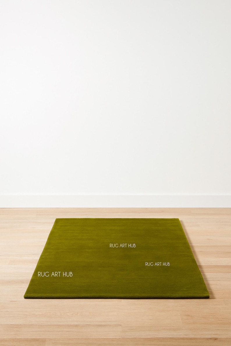 May include: A rectangular, olive green rug with a textured surface. The rug is placed on a light-colored wooden floor against a white wall. The text "RUG ART HUB" is printed on the rug.