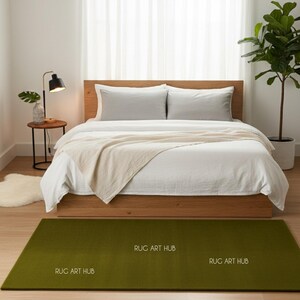 May include: A bedroom scene featuring a bed with white bedding, a wooden frame, and two gray pillows. A green rectangular rug with the text "RUG ART HUB" is on the floor. A small side table with a lamp and a plant is next to the bed.