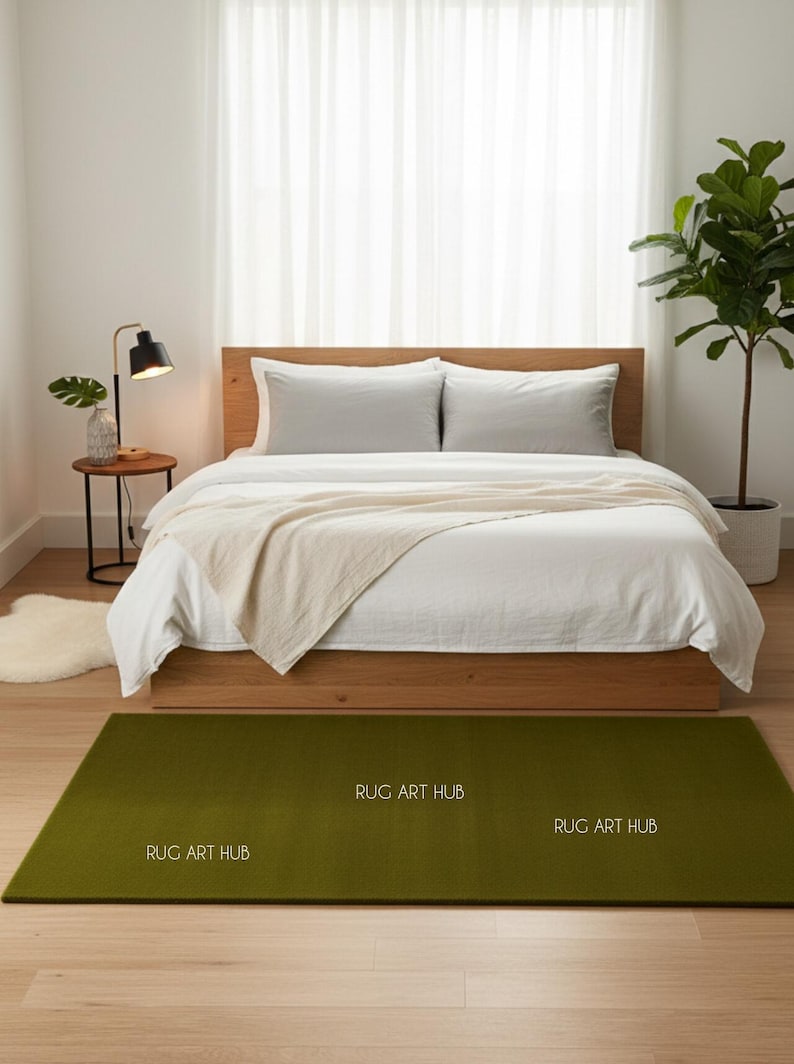 May include: A bedroom scene featuring a bed with white bedding, a wooden frame, and two gray pillows. A green rectangular rug with the text "RUG ART HUB" is on the floor. A small side table with a lamp and a plant is next to the bed.