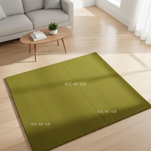 May include: A large, rectangular olive green rug with a textured surface. The rug is placed on a light wood floor in a living room setting. The words "RUG ART HUB" are printed on the rug.