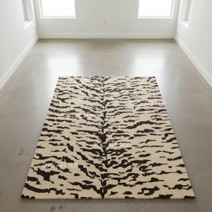 Puede incluir: Una alfombra rectangular con un diseño de rayas de tigre. La alfombra presenta un fondo color crema con rayas marrones oscuras. Está colocada sobre un suelo de hormigón gris en una habitación con paredes y ventanas blancas.