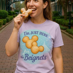 May include: A light purple t-shirt with the text "I'm JUST HERE for the Beignets" and a graphic of Mickey Mouse-shaped beignets. The person is eating a beignet. The shirt is made of a soft material.