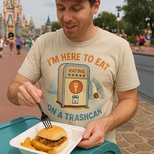 May include: A person wearing a tan t-shirt with the text "I'M HERE TO EAT ON A TRASHCAN" and a graphic of a trash can with a fork. The person is holding a burger and fries. A gray cap is worn.