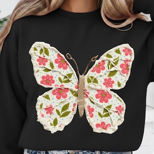 Könnte beinhalten: Schwarzes Sweatshirt mit einem großen Schmetterlingsdesign. Der Schmetterling hat cremefarbene Flügel mit einem Blumenmuster in Rosa und Grün, akzentuiert mit blauen Punkten. Der Körper des Schmetterlings ist ein strukturiertes braunes Material.