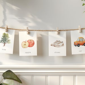 May include: Four watercolor illustrations of everyday objects are displayed on a rope with wooden clips. The images include a tree, an apple, a pair of shoes, and a car. Each image has a corresponding word written below it.