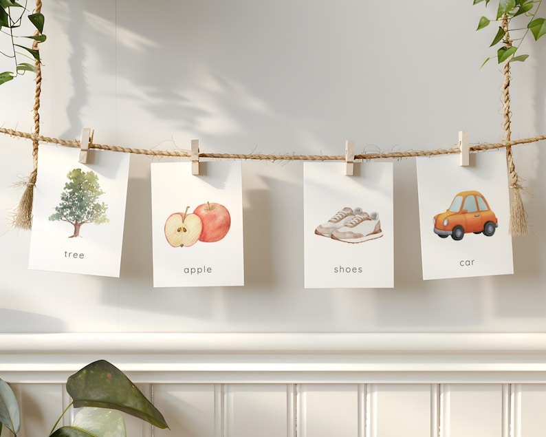 May include: Four watercolor illustrations of everyday objects are displayed on a rope with wooden clips. The images include a tree, an apple, a pair of shoes, and a car. Each image has a corresponding word written below it.