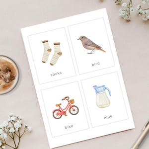 May include: A card with four watercolor illustrations: a pair of socks, a bird, a red bike, and a milk pitcher. Each image has a word label beneath it: "socks", "bird", "bike", and "milk". The card is on a light-colored surface.
