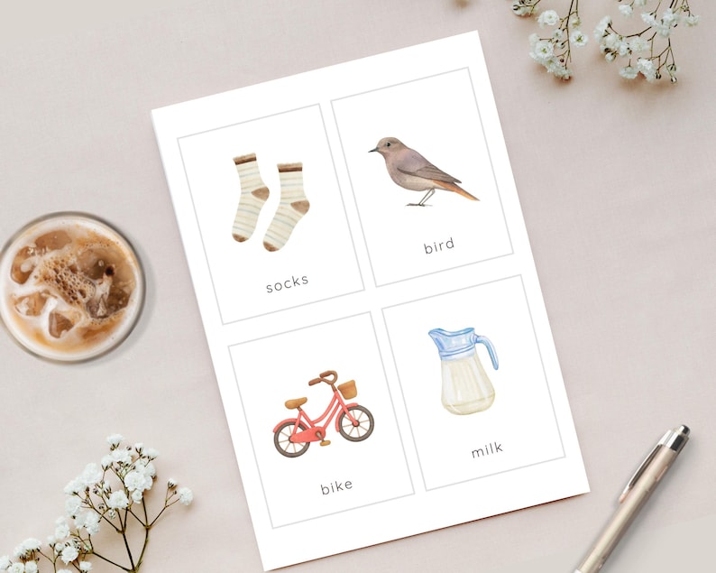 May include: A card with four watercolor illustrations: a pair of socks, a bird, a red bike, and a milk pitcher. Each image has a word label beneath it: "socks", "bird", "bike", and "milk". The card is on a light-colored surface.