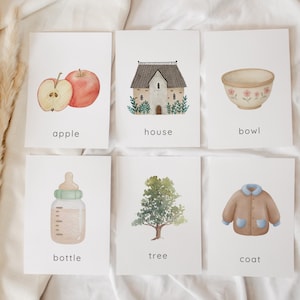 Puede incluir: Seis tarjetas didácticas de acuarela con ilustraciones de una manzana, una casa, un cuenco, un biberón, un árbol y un abrigo. Cada tarjeta tiene el nombre del objeto debajo. La manzana es roja y cortada. El abrigo es marrón con detalles azules.