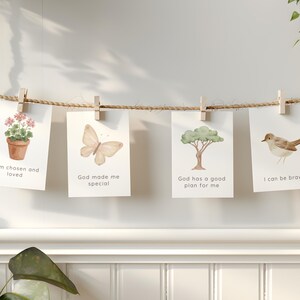 May include: Four rectangular white cards hanging from a horizontal jute rope with wooden clips. The cards feature watercolor illustrations of a potted plant, a butterfly, a tree, and a bird, with the text: I am chosen and loved, God made me special, God has a good plan for me, and I can be brave.