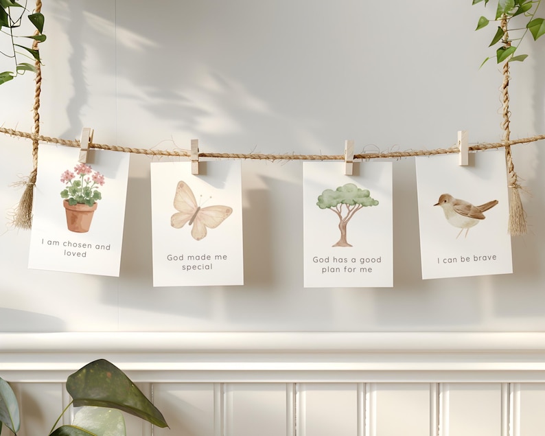 May include: Four rectangular white cards hanging from a horizontal jute rope with wooden clips. The cards feature watercolor illustrations of a potted plant, a butterfly, a tree, and a bird, with the text: I am chosen and loved, God made me special, God has a good plan for me, and I can be brave.