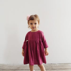 May include: A young child wearing a burgundy linen dress with three-quarter sleeves and white shoes. A pink flower hair accessory is pinned to the child's hair. The dress has a gathered skirt and a simple, round neckline.