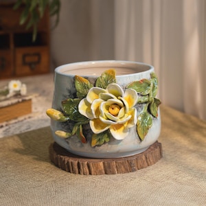 May include: A ceramic planter with a light blue and gray finish, adorned with a 3D yellow rose and green leaves. The flower pot sits on a round, wooden coaster. The design is detailed and artistic, suitable for indoor or outdoor use.