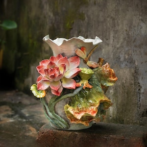 以下が含まれることがあります： 詳細な蓮の花のデザインが施されたセラミック花瓶。花瓶は、フリル付きの縁が付いた明るい灰色のベースが特徴で、赤、ピンク、黄色の色合いの大きな立体的な蓮の花で飾られています。緑の葉とつぼみが花のテーマを補完しています。