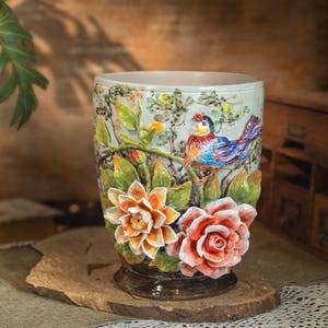 May include: A ceramic vase with a detailed relief design. The vase features a colourful bird perched on a branch, surrounded by sculpted flowers in shades of orange and red. The background is a light green with floral accents.