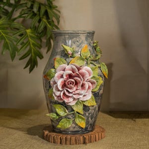 May include: A decorative ceramic vase with a large, detailed rose design. The vase is gray with a marble-like texture, and the rose is pink and white with green leaves. The vase sits on a wooden base, creating a floral theme.