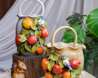 Puede incluir: Dos cestas de cerámica con asas, adornadas con frutas rojas y naranjas esculpidas, hojas verdes y pequeños pájaros azules. Una cesta es gris, la otra es marrón. Una pequeña figura de ardilla está cerca.