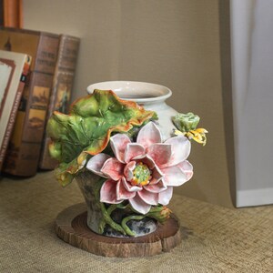 Könnte beinhalten: Eine Keramikvase mit Lotusblüten-Design. Die cremefarbene Vase hat eine große rosa-weiße Lotusblüte, grüne Blätter und gelbe Akzente. Sie steht auf einem runden Holzsockel. Der Stil ist dekorativ und ornamental.