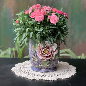 May include: A ceramic planter with a glossy purple and blue marbled finish, featuring a large, three-dimensional sculpted flower on the front. The planter is filled with a dense cluster of vibrant pink carnations and green foliage.