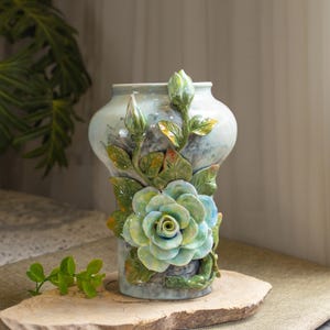 May include: A ceramic vase with a pale blue and grey mottled finish. The vase features a large, three-dimensional, light green and blue rose and two rose buds. The leaves are green with yellow and red accents. The vase is displayed on a wooden slab.